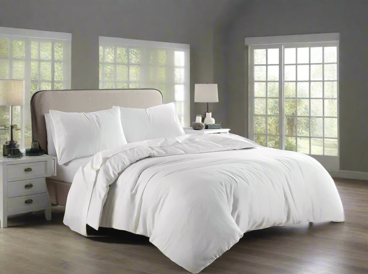White bedding set on a bed in a bright bedroom with large windows.